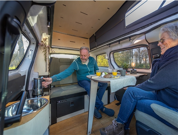 Met de draaibare keuken van de Outbase ID.Buzz kun je zowel binnen als buiten koken. De afbeelding laat een koppel zien wie binnen de camper zitten te ontbijten en koffie zetten op de draaibare keuken. 